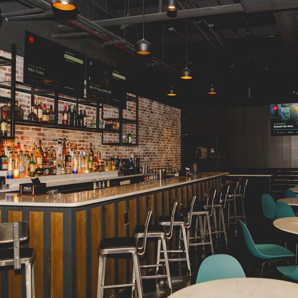 A dimly lit bar area with a brick wall, liquor bottles, bar stools, and teal chairs.