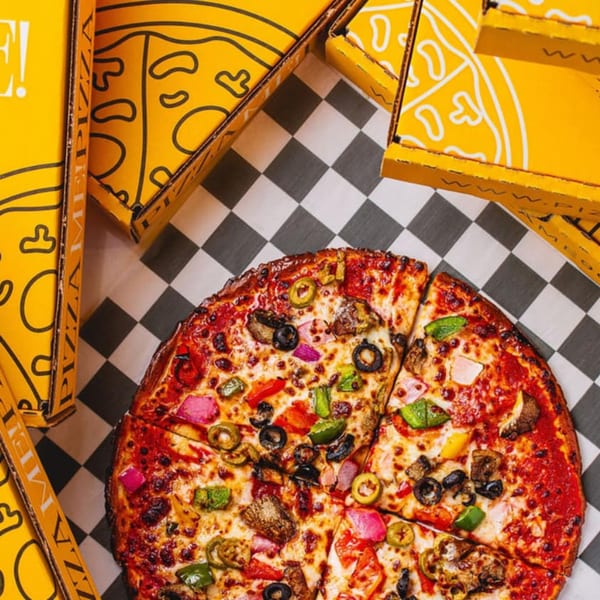 A PizzaMe! supreme pizza with various toppings on a checkered tablecloth, surrounded by yellow pizza boxes.