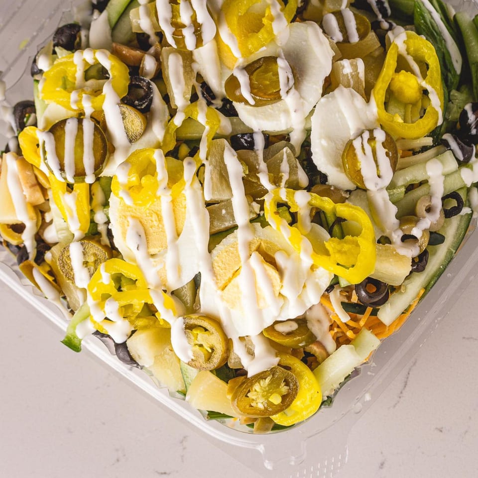 A close-up of a vibrant salad with yellow peppers, black olives, and white dressing in a clear container.