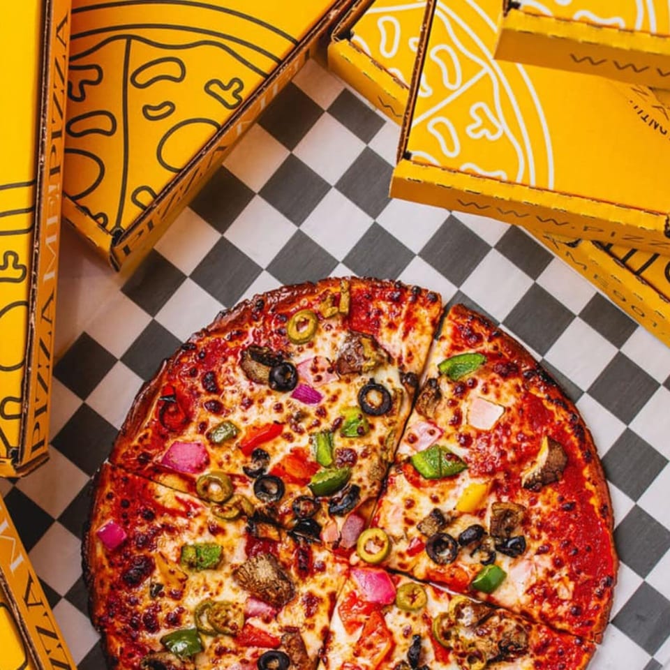 A PizzaMe! pizza with various toppings sits on a checkered surface surrounded by yellow pizza boxes.