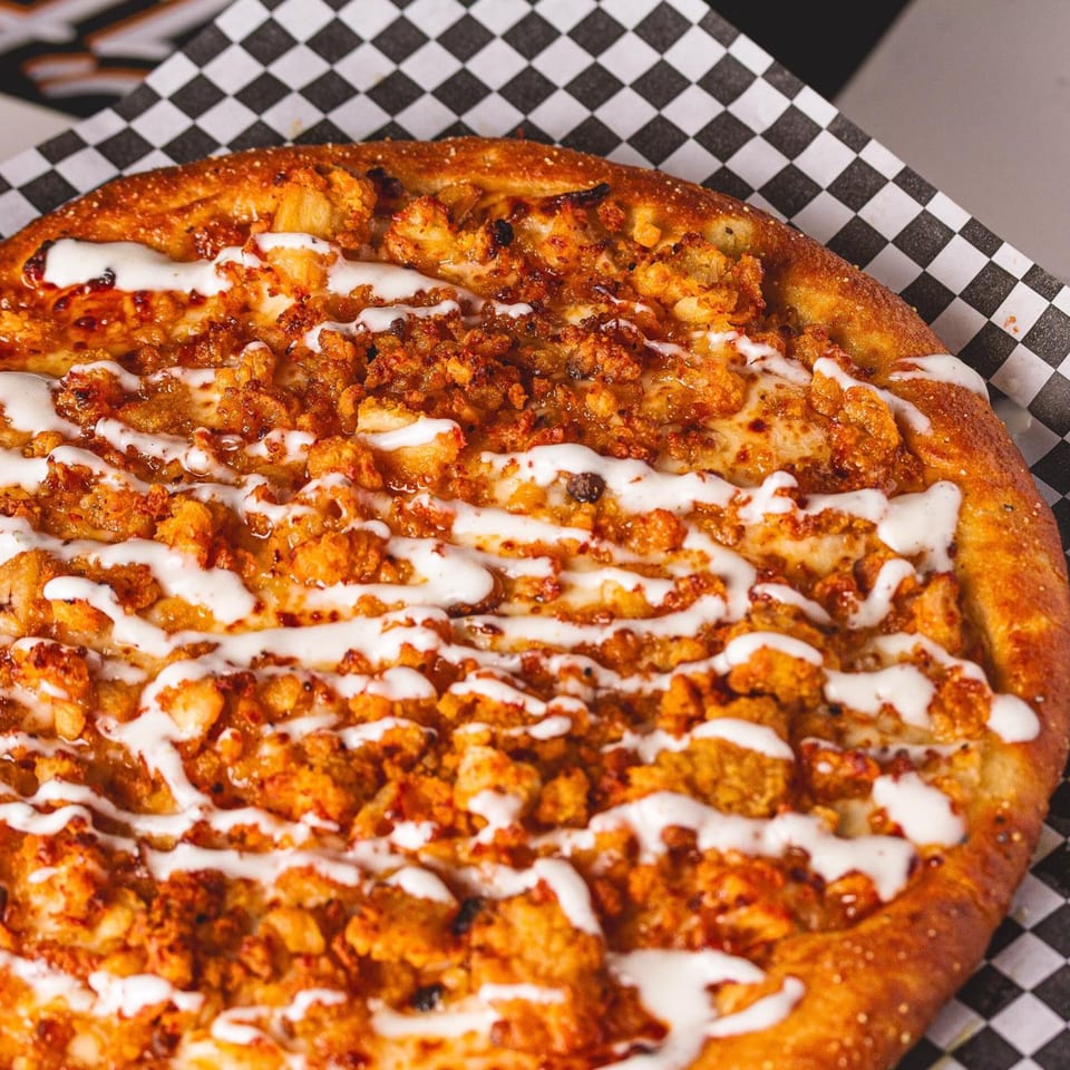 Close-up of a PizzaMe! buffalo chicken pizza with white sauce drizzle on checkered paper.