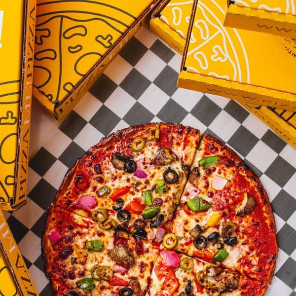 A PizzaMe! supreme pizza with various toppings on a checkered tablecloth, surrounded by yellow pizza boxes.