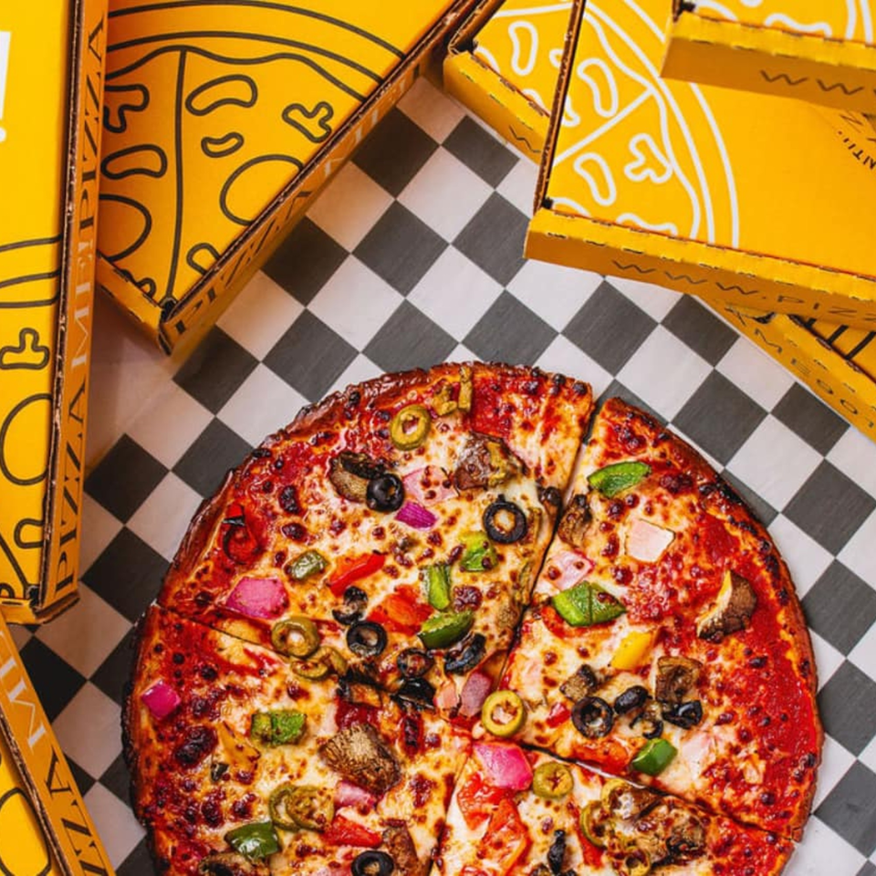 A PizzaMe! supreme pizza with various toppings on a checkered tablecloth, surrounded by yellow pizza boxes.