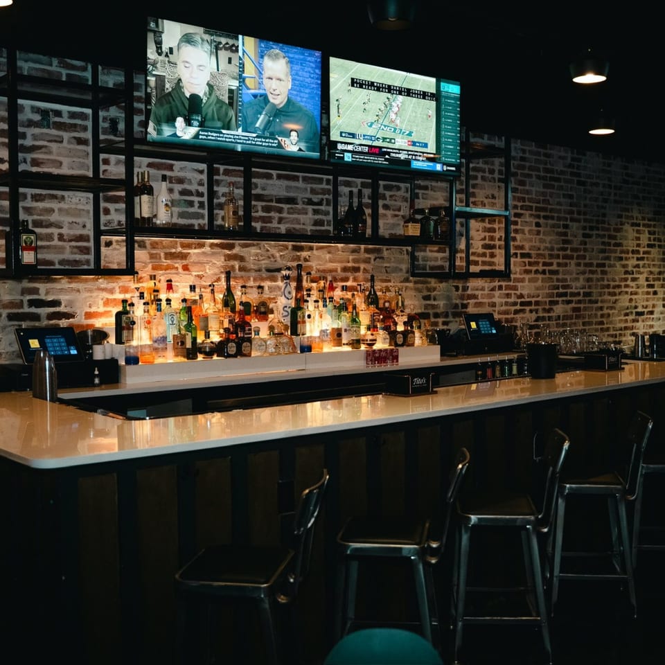 A dimly lit bar with a brick wall, liquor bottles, and three TVs showing sports.